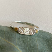 A polished 925 sterling silver band ring with a central plaque featuring the engraved letters 'XXX' flanked by two small brass dots on either side, placed on a leaf.