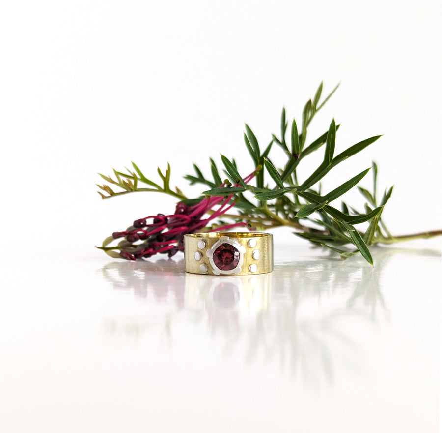 A silver ring with a round garnet in a raised bezel setting, accompanied by a yellow brass band.