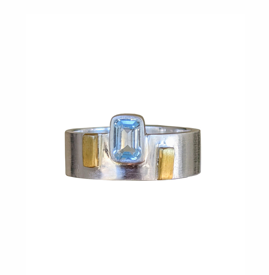 A sterling silver ring with a baguette cut blue topaz centerpiece and yellow brass accents on the band.